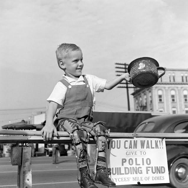 La imagen muestra a un niño con aparatos ortopédicos en las piernas,  sentado en la calle, sonriendo y sosteniendo una cacerola junto a un cartel que  indica "tu puedes caminar, contribuye con monedas para la polio".  Fotografía de Martha Homes, 1940.