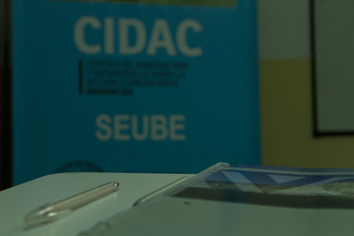 La imagen muestra en primer plano un cuaderno y una lapicera y, de fondo, un banner del CIDAC.