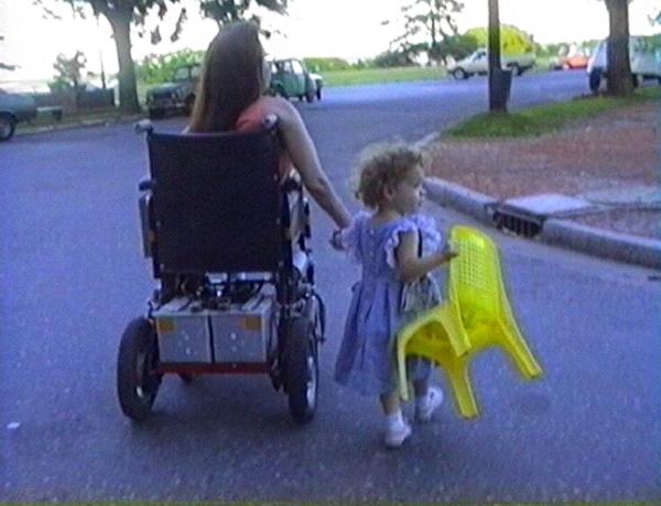 La imagen muestra a una mujer en silla de ruedas que pasea con su hija. La niña lleva en la mano una silla de plástico.